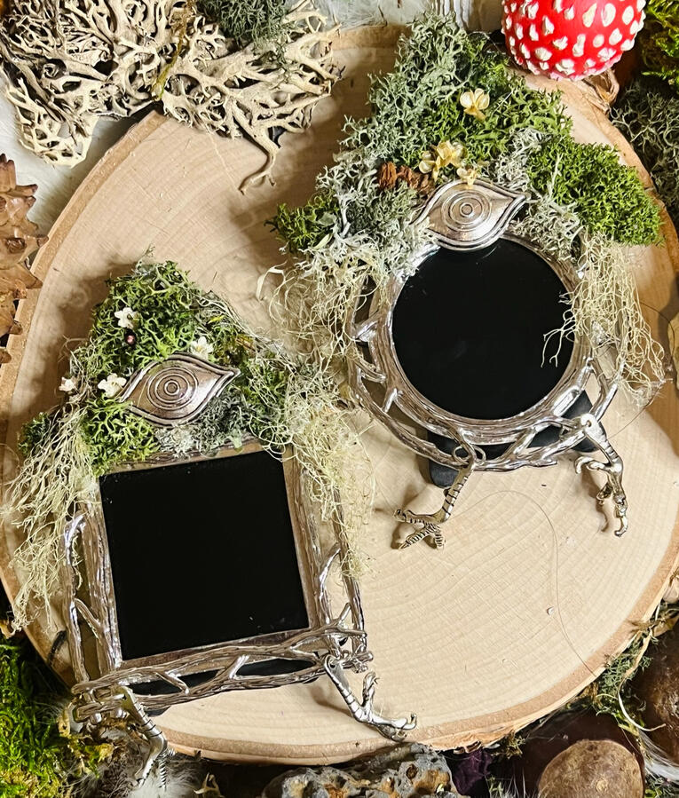 mini scrying mirrors reminiscent of Baba Yaga's hut, with a mossy, flowered roof containing a silver, metal eye atop a silver, metal frame with a branch motif, finished with silver metal chicken's feet